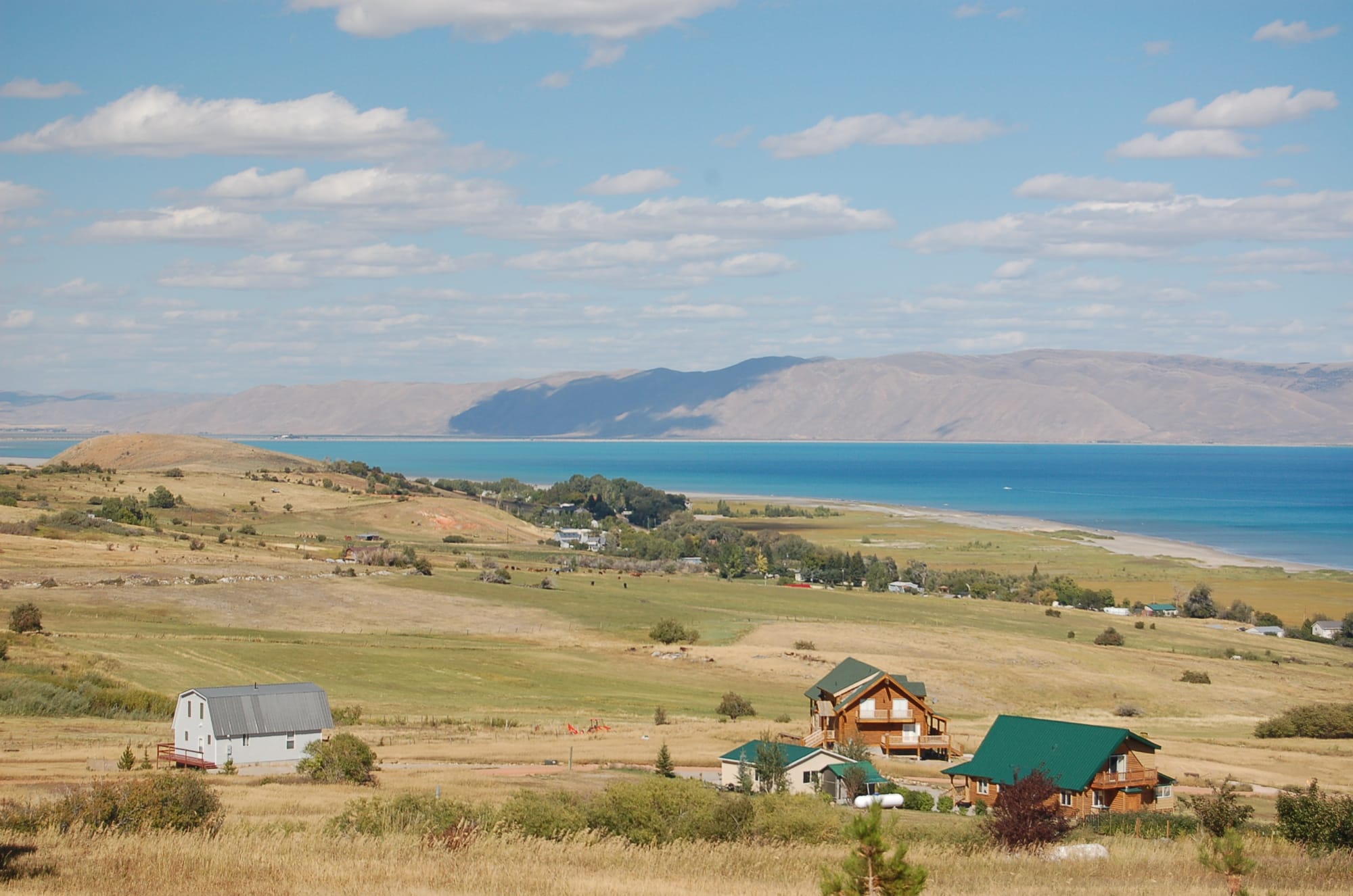 Hay fields, a barn and scattered houses on the valley floor, with the turquoise water of Bear Lake and the Bear River Range beyond